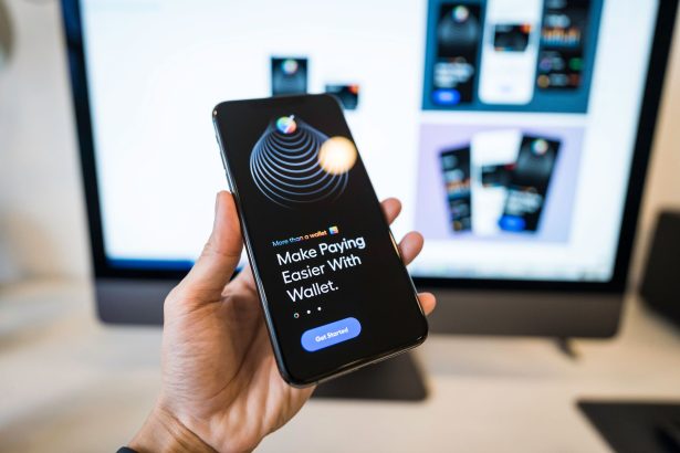 Pessoa segura um celular com um aplicativo de carteira digital aberto, diante de um monitor desfocado.