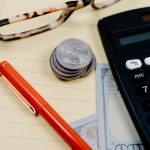 Calculadora, moedas e óculos sobre um bloco de anotações em uma mesa de trabalho financeiro