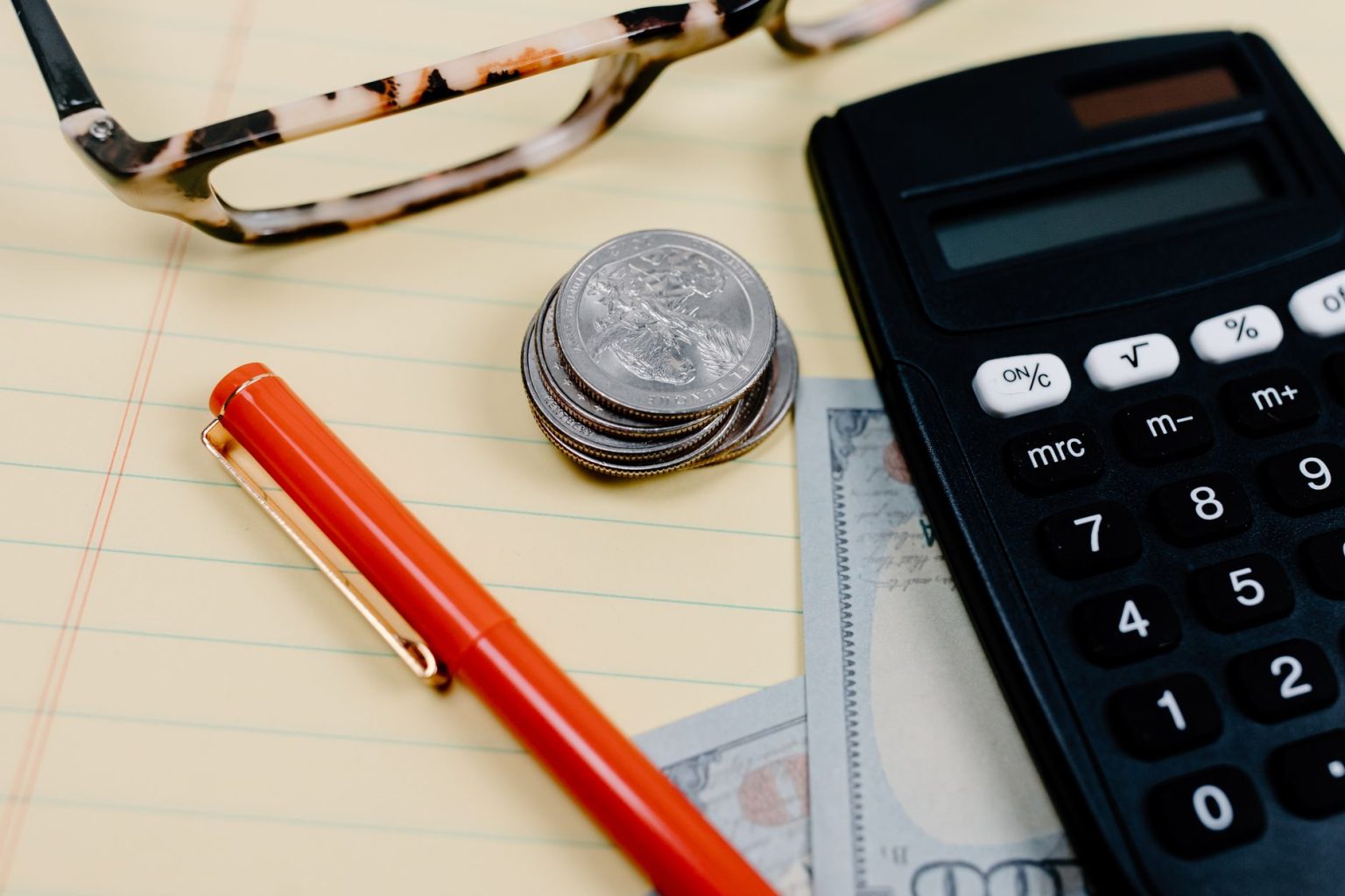 Calculadora, moedas e óculos sobre um bloco de anotações em uma mesa de trabalho financeiro